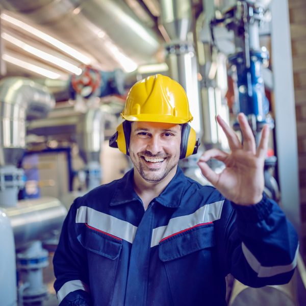 Smiling satisfied blue collar worker in working clothes, with helmet and hardhat standing in factory and showing okay sign.